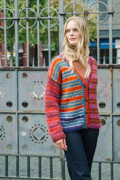 a woman standing in front of a gate wearing a multicolored cardigan sweater