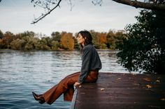 a woman sitting on the edge of a dock next to a body of water with trees in the background