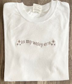 Embroidered in my wifey era  comfort color tee shirt,  * custom in my era list in personalization option along with your thread color choice ( one color please)!  Tee Shirt description : *Tee is true to size ,preshrunk/Relaxed fitting and is made from 100% ring spun cotton.   -Size Chart is listed in Images!  - Size up for oversize look! THREAD COLORS: white, black, charcoal grey, silver grey, sand, golden pecan, taupe/image, brown,ivory, dusty blue, golden yellow, mauve, dark purple, moss green Customizable Cotton Tops For Anniversary, In My Bride Era, Wife Apparel, Engagement Shirt, Wifey Shirt, Engaged Shirts, Bride Shirt, Bride Shirts, Comfort Colors Tee