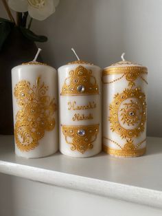 three white candles with gold embellishments are sitting on a shelf next to a flower