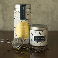 two tins with tea leaves and a strainer next to them on a table