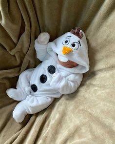 a baby in a snowman costume laying on a bed with a brown blanket behind it