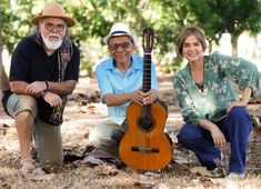 two men and a woman are sitting in the woods with an acoustic guitar on their lap