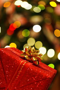 a red gift box with a gold bow on it sitting in front of a christmas tree