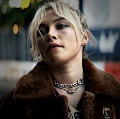 a woman with blonde hair wearing a brown coat and silver necklace looking off into the distance