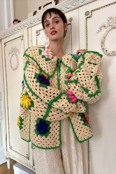 a woman standing in front of a white cabinet wearing a crocheted jacket with flowers on it