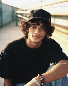 a young man with curly hair wearing a black shirt and cap sitting on a bench