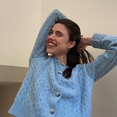 a woman in a blue sweater is smiling and holding her hands behind her head with one hand