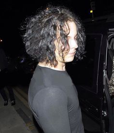 a close up of a person with curly hair near a car at night, looking off to the side