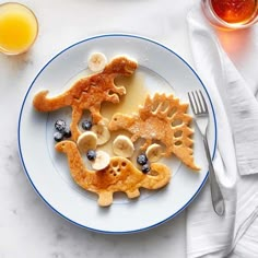 a white plate topped with pancakes covered in banana slices and blueberries next to a glass of orange juice