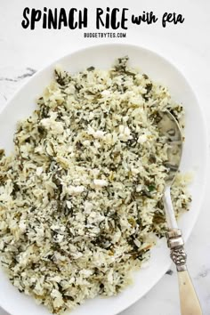 a white plate topped with rice and spinach next to a silver spoon on a marble surface