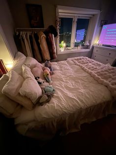 an unmade bed with stuffed animals on it in front of a window at night