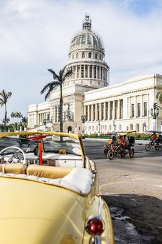 an old yellow car parked in front of a large building with a dome on top