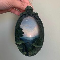 a hand is holding an oval painting with trees on it