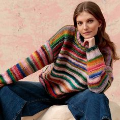 a woman sitting on top of a wooden bench wearing a multicolored sweater and jeans