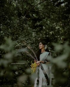 a woman in a white sari holding flowers and looking up into the sky with trees behind her