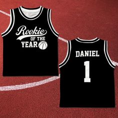 two black basketball jerseys with white lettering on the front and back, sitting on a red court