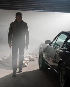 a man standing next to a parked car in a garage with dust on the ground