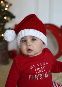 a baby wearing a red shirt and white hat sitting in front of a christmas tree