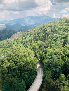 an aerial view of a road in the middle of a forest with mountains in the background