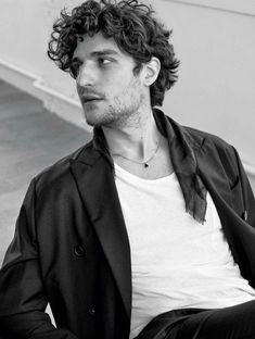 a man with curly hair is sitting on the ground wearing a white t - shirt and black blazer