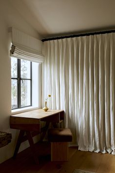 a desk and chair in front of a window with white drapes on the windowsill