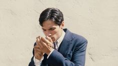 a man in a suit and tie praying with his hands folded over his chest, against a white wall