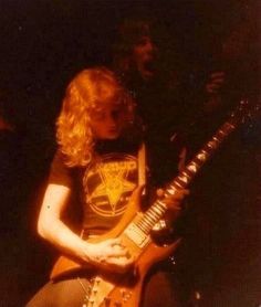 a man playing an electric guitar in front of another person with long hair and wearing a t - shirt