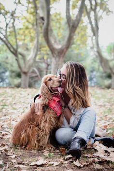 a woman sitting on the ground with her dog in front of trees and fallen leaves