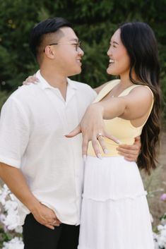 a man and woman standing next to each other with their arms around each other in front of flowers