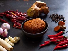 various vegetables and spices are arranged on a black counter top, including red peppers, garlic, ginger, tumerics, and other ingredients