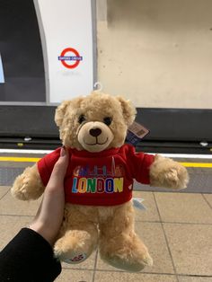 a person holding a brown teddy bear wearing a london t - shirt at an airport