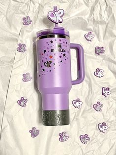 a purple travel mug sitting on top of a white sheet with hearts and butterflies around it