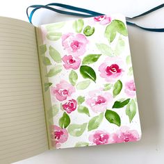 an open notebook with watercolor flowers and green leaves on the cover, next to a blue ribbon