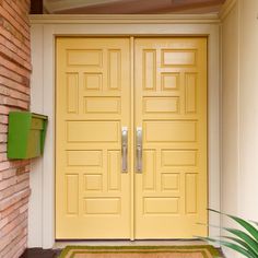 a yellow door with two green planters on the side and a red brick wall behind it