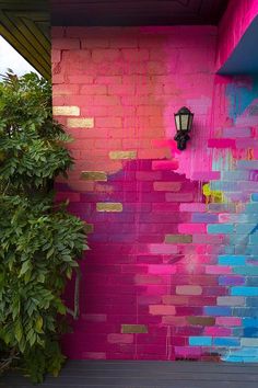 a pink and blue painted brick wall next to a green bush with a lamp on it