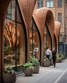 two people are walking down the sidewalk in front of large windows with planters on them