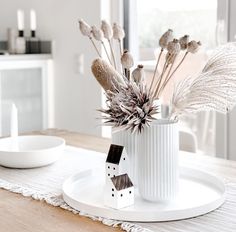 a white vase filled with dried plants on top of a wooden table next to a plate