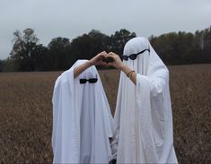 two women dressed in white are making heart shapes with their hands while standing in the middle of a field