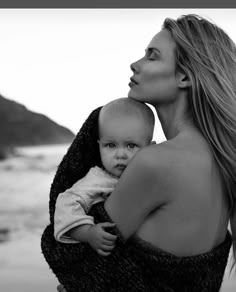 a woman holding a baby in her arms on the beach with mountains in the background