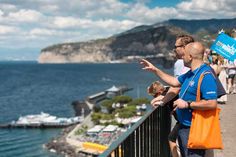 two men are pointing at something while standing on a balcony overlooking the water and mountains