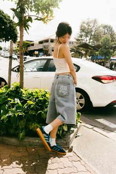 a woman standing on the sidewalk in front of a white car and some bushes with her feet up