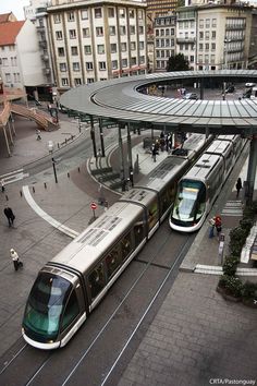 two trains passing each other on the tracks in front of an overpass with people walking around