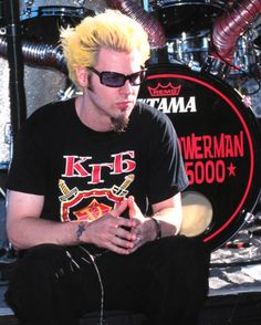 a man with yellow hair and sunglasses sitting in front of a drum kit on stage