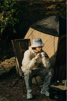 a man sitting in a chair next to a tent and holding a cup with his hand