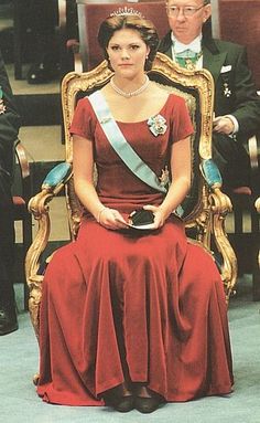 a woman in a red dress is sitting on a gold chair with other people behind her