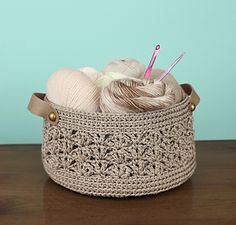 a crocheted basket with yarn and knitting needles in it on a wooden table