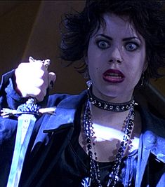 a woman with dark hair and blue eyes holds a knife in her hand while wearing an elaborate necklace