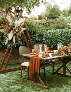 an outdoor dining table set with orange and white flowers on it, surrounded by greenery