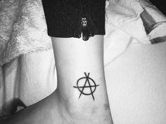 a small tattoo on the ankle of a person with a cross and anarchy symbol in black ink